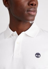 Balta polo krekla ar struktūru. Tam ir trīs pogu atvērums un tumši zils izšūts koka logos kreisajā krūtī.