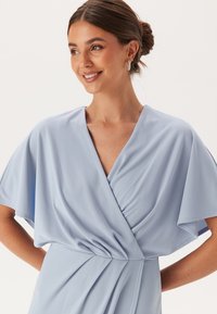 Jasnoniebieska sukienka z dekoltem w kształcie litery V, drapowanymi krótkimi rękawami i marszczoną talią, wykonana z miękkiego materiału, o gładkiej teksturze i luźnym kroju.