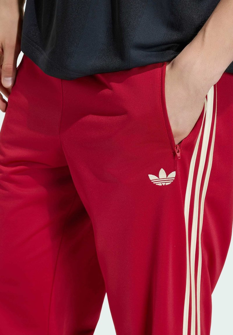 Person iført røde Adidas træningsbukser med hvide striber og en sort skjorte, hånd i lynlåslomme på højre side.