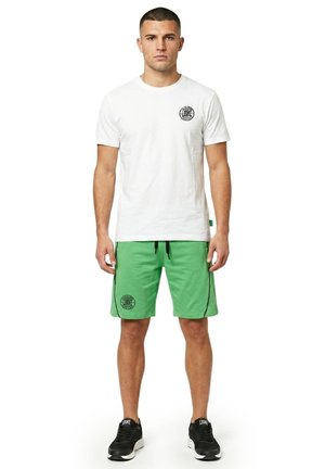 Un homme présente un tee-shirt blanc à logo, un short vert et des baskets noires sur un fond blanc uni.