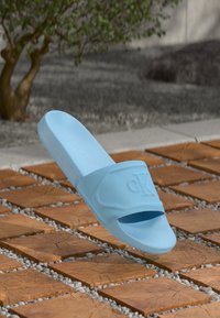 Ljust blå gummisandaler med en utskuren rem och präglad logotyp. Har en platt sula och slät yta, svävande ovanför en kaklad yta.