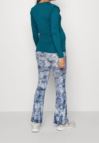 Teal långärmad topp med rynkade axeldetaljer, tillsammans med blå batikmönstrade utsvängda byxor. Modellen bär vita sneakers.