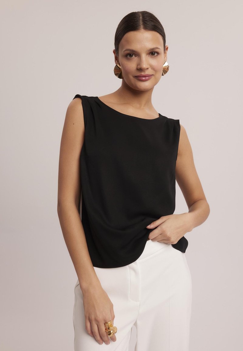 Czarna bezrękawnikowa bluzka z okrągłym dekoltem, drapowany design i miękka tkanina; w połączeniu z białymi spodniami i złotymi, wyrazistymi kolczykami.