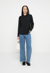 Fekete hosszú ujjú ing gallérral, széles szárú kék farmerrel, fekete cipővel és kis fehér kézitáskával párosítva. Egyszerű, klasszikus kialakítás.