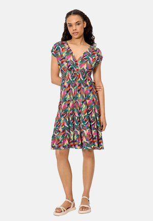 Mujer con un vestido hasta la rodilla de estampado multicolor con hojas, manga corta y sandalias blancas, de pie contra un fondo liso.