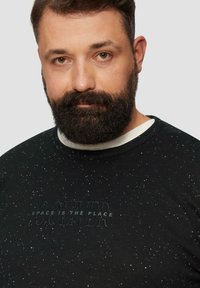 Schwarzer Pullover mit weißen Sprenkeln, ausgestattet mit einem strukturierten Grafikelement, das in erhabener Schrift "SPACE IS THE PLACE" präsentiert.