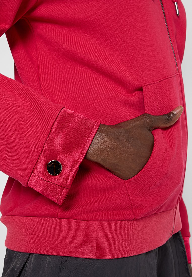 Una mano di carnagione scura con il pollice alzato nel taschino di una felpa rossa con zip, con polsini in velluto e dettagli in bottone nero.