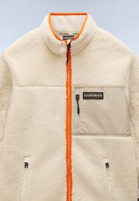 Cream fleece jacket with orange accents, zip closure, high collar, and beige pocket. Textured material and branded patch detail on chest.