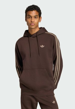Młody mężczyzna w ciemnobrązowej bluzie z kapturem marki Adidas i pasujących spodniach z beżowymi paskami, zwrócony przodem na jednolitym jasnym tle.
