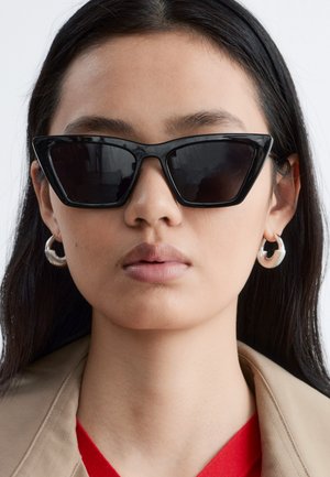 Femme aux longs cheveux foncés portant des lunettes de soleil noires angulaires, des boucles d'oreilles anneaux en argent, une veste beige et un haut rouge sur un fond neutre.