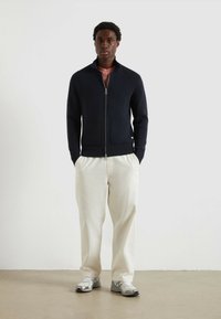 Maglione a zip di colore blu navy con design tessurizzato, due tasche frontali, abbinato a pantaloni crema e sneakers grigie. Aspetto semplice e moderno.