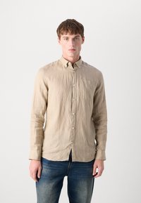 Camisa bege de manga longa abotoada em tecido leve e texturizado; possui um bolso no peito e colarinho padrão. Usada com jeans azuis.