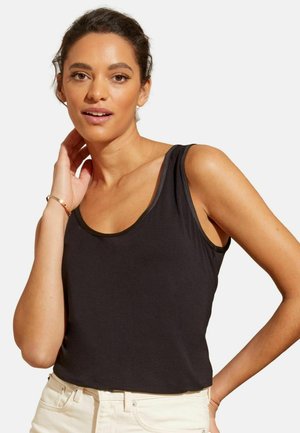 Femeie cu păr închis la culoare, purtând un top negru fără mâneci și pantaloni deschiși la culoare, atingându-și gâtul cu o mână, pe un fundal alb simplu.