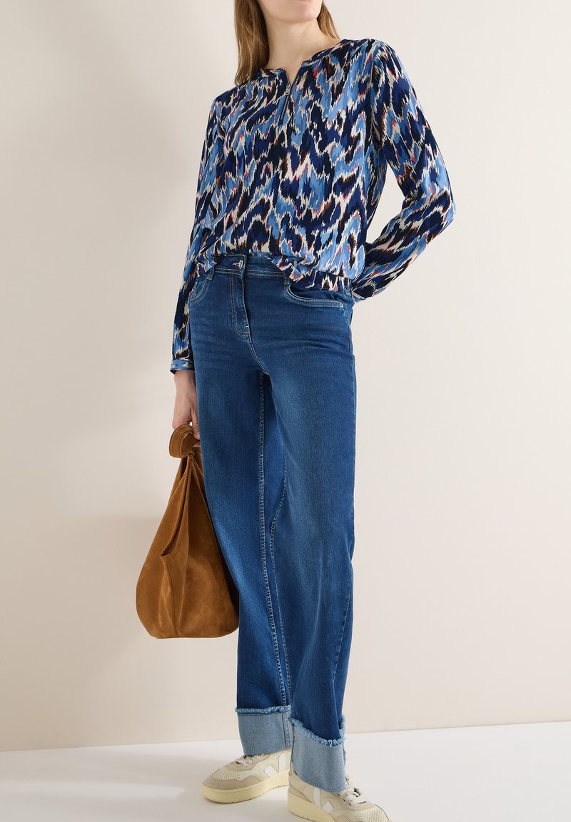 Femme portant un chemisier à manches longues bleu à motifs, un jean large à revers bleu, des baskets beiges, et tenant un sac à main en daim marron.