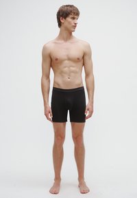 Juodi bokso šortai, su elastine juosmens juosta, prigludusiu dizainu, minkšta medžiaga, trumpais kojų ilgiais, modelis basomis stovintis priešais paprastą foną.