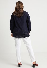 Blouse bleu marine avec des détails en œillets floraux et un ourlet festonné, associée à un pantalon blanc. La tenue présente des manches longues et une coupe décontractée.