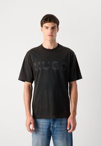 Juodas medvilnės marškinėliai su laisva siluetu, ant krūtinės yra toninis "HUGO" logotipas, trumpomis rankovėmis ir apvalia iškirpte. Mėlyni džinsai.