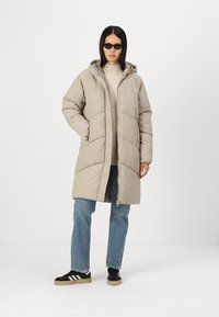 Casaco acolchoado bege com capuz, apresenta um design acolchoado, fecho frontal e dois bolsos laterais. Usado sobre um suéter leve e jeans.