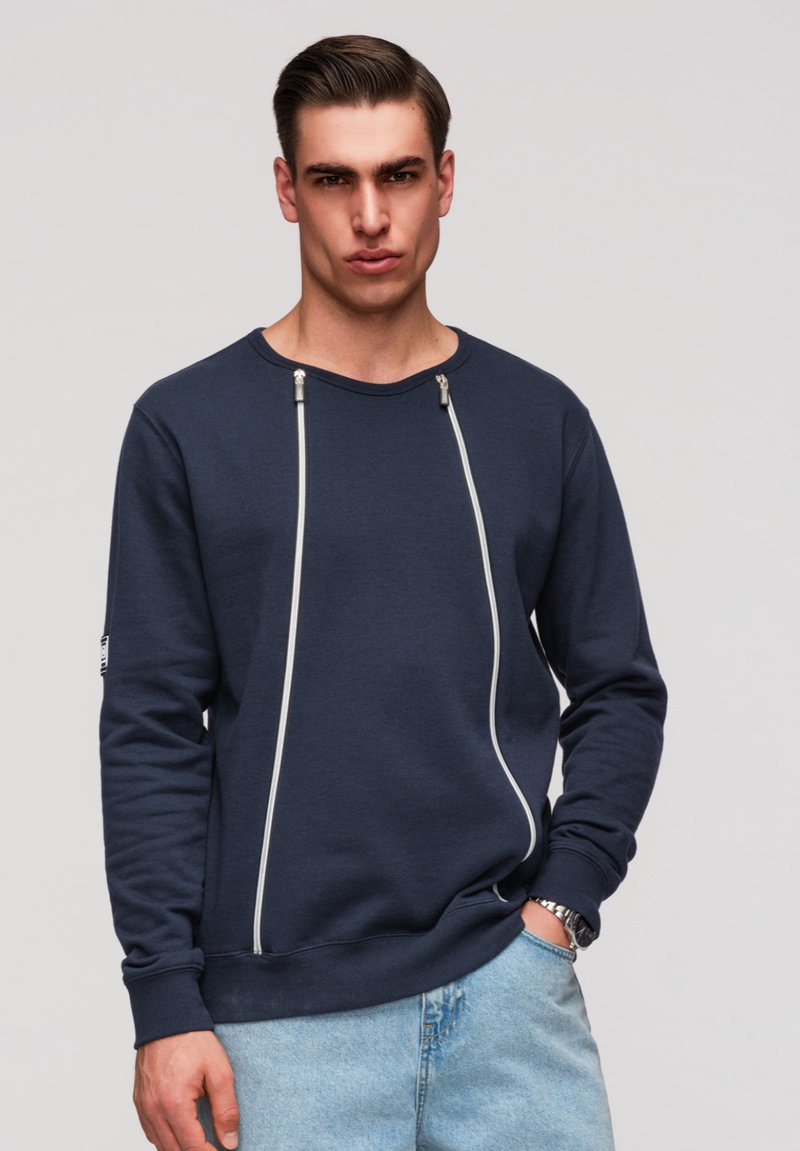 Homme portant un sweat-shirt bleu marine avec deux fermetures éclair verticales et un jean bleu clair, debout avec une main dans la poche.