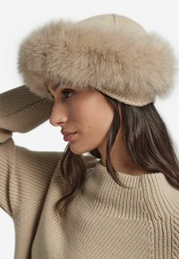 Beige stickad hatt med bred kant i konstpäls. Texturen är mjuk och formen rundad, vilket säkerställer en bekväm passform runt huvudet.