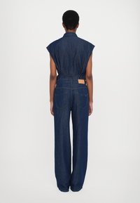 Macacão de ganga com mangas curtas e largas, cintura com cinto e pernas largas de corte solto. Azul escuro, com bolsos traseiros e um pequeno detalhe de etiqueta.