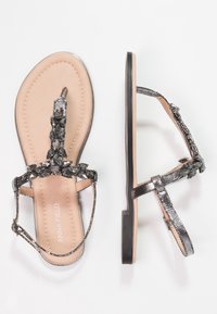 Litteet thong-sandaalit, joissa on metallinen musta hihna, koristeltuna tummilla jalokivillä, ja beige pohja. Pintakuvioinnit ylähihnassa.