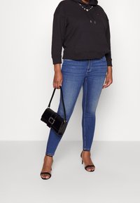 Sudadera negra con capucha y bolsillo, jeans skinny azules y sandalias de tacón negras. Sosteniendo un pequeño bolso negro con una hebilla plateada.