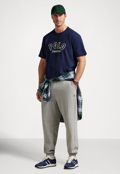 Moški z zeleno kapo, mornarsko modro majico "Polo", sivimi športnimi hlačami, modrimi supergami in karo srajco, vezano okoli pasu, stoji z rokami v žepih.