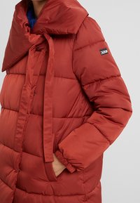 Cappotto imbottito rosso con grande colletto, chiusura anteriore legata, polsini elasticizzati e tasche laterali. Presenta una toppa con logo sulla manica sinistra.