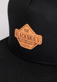 Mustat hattu, jossa on teksturoitu ruskea nahkapatchi, jossa on sanat "BLACKSKIES PREMIUM SUPPLIES" ja koristeellisia yksityiskohtia.