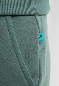 Zelené bavlněné kalhoty s ribbovaným pasem, bočními kapsami a malou tyrkysovou etiketou. Materiál má hladký, strukturovaný povrch.