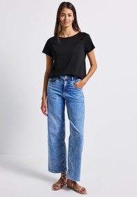 Zwarte kortekorte mouw top gecombineerd met lichtblauwe wijde jeans. Sandalen zijn metallic met meerdere banden. Effen achtergrond.