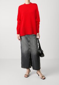 Pull en maille rouge à texture côtelée, associé à une jupe longue en jean avec une fente à l'avant, combiné avec des sandales à talons noires et un sac à main.