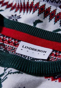 Pulôver de malha com uma base branca e padrões em vermelho e verde, colarinho canelado verde e uma faixa de destaque vermelha. Inclui um rótulo "Lindbergh White".