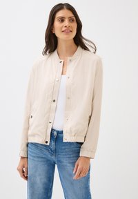 Lichtbeige bomberjack met een ronde hals, drukknoopjes en geribbelde manchetten. Draag het over een witte top en combineer het met blauwe jeans.