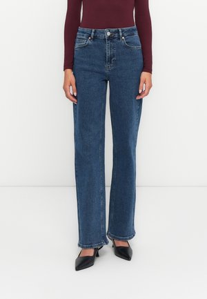Woman wearing high-waist straight-leg blue jeans, burgundy long-sleeve top, and black pointed-toe heels standing on white floor.