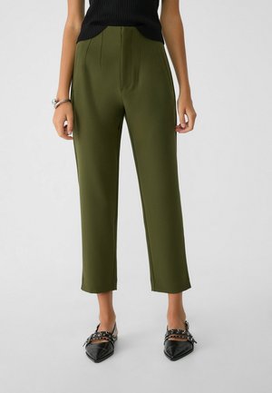 Personne portant un pantalon court vert olive, un haut noir sans manches, des chaussures noires pointues à boucles, debout devant un fond uni.