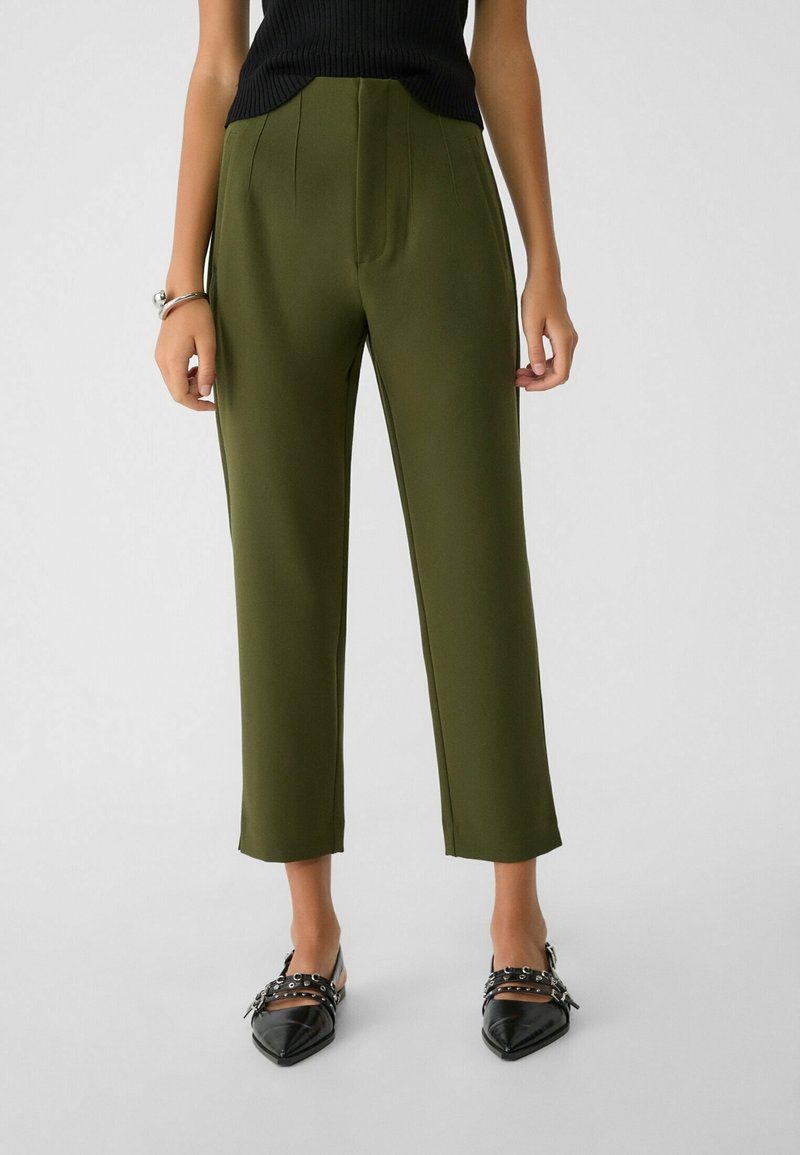 Personne portant un pantalon court vert olive, un haut noir sans manches, des chaussures noires pointues à boucles, debout devant un fond uni.