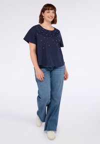 Donna sorridente che cammina, indossando una maglietta blu navy a maniche corte con dettagli di perle, jeans a gamba larga con orli sfrangiati e sneaker bianche.