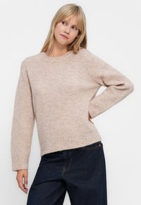 Ροζ πλεκτό πουλόβερ με στρογγυλή λαιμόκοψη και μακριά μανίκια, προσφέροντας ribbed στο τελείωμα και πλευρικές σχισμές για επιπλέον λεπτομέρεια. Φορεμένο με σκούρο τζιν.