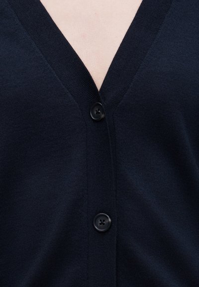 Κοντινή λήψη σκούρου μπλε ζακέτας με δύο μαύρα κουμπιά κλειστά, που δείχνει V-λαιμόκοψη και υφή πλεκτού υφάσματος.