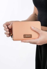 Peach-colored wallet with a smooth, matte finish, zip closure, and a brown leather label. Handheld against a white background.