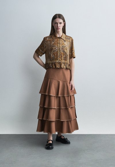 Γυναίκα που φοράει καφέ μπλούζα με μοτίβο και κοντά μανίκια, και καφέ φούστα με στρώσεις και διακοσμητικές πινελιές με τρουκς, στέκεται μπροστά σε γκρι απλή τοίχο.