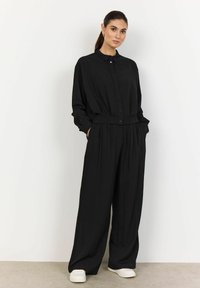 Jumpsuit nero con bottoni, maniche lunghe e vita cinchata. Realizzato in un tessuto leggero eTexture, con pantaloni a gamba larga.