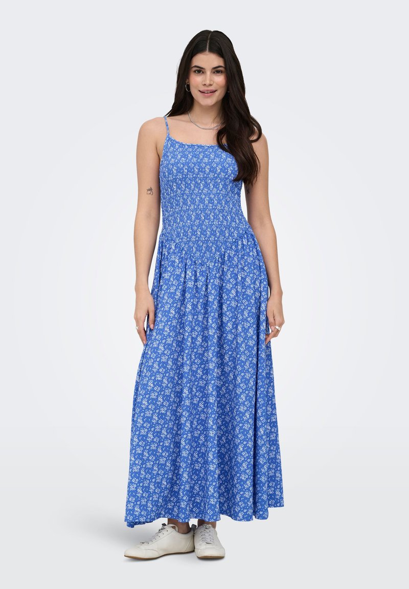 Femme portant une robe longue sans manches bleue à fleurs avec des fines bretelles et des baskets blanches, debout devant un fond uni.