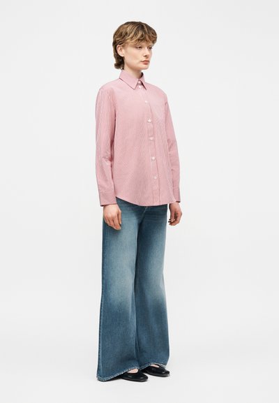 Άτομο που στέκεται φορώντας ροζ ριγέ πουκάμισο με κουμπιά, φαρδιά ξεβάμμένα μπλε τζιν και μαύρα φλατ παπούτσια, μπροστά σε απλό φόντο.