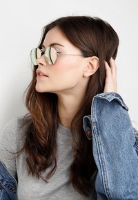 Runde, sølvfarvede solbriller med spejl-linser. Båret af en person med langt, bølget hår, iført en grå top og en denimjakke.