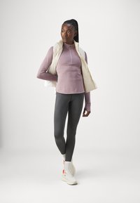 Gilet en créme léger au-dessus d'un haut mauve à manches longues ajusté, associé à des leggings gris foncé. La modèle porte des baskets blanches avec des accents orange.
