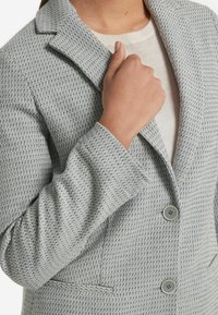 Grå vävd blazer med en snibbkrage, två knappar och en bröstficka. Materialet verkar vara vävt med ett subtilt mönster.