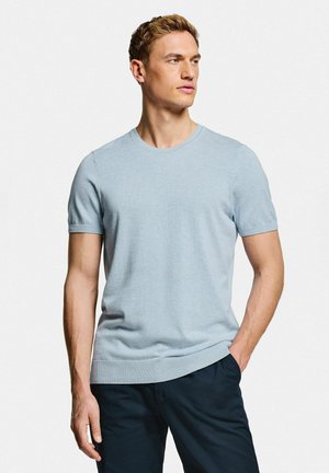 Jeune homme portant une chemise en tricot à manches courtes bleu clair et un pantalon foncé, debout avec une main dans la poche, regardant vers sa gauche.